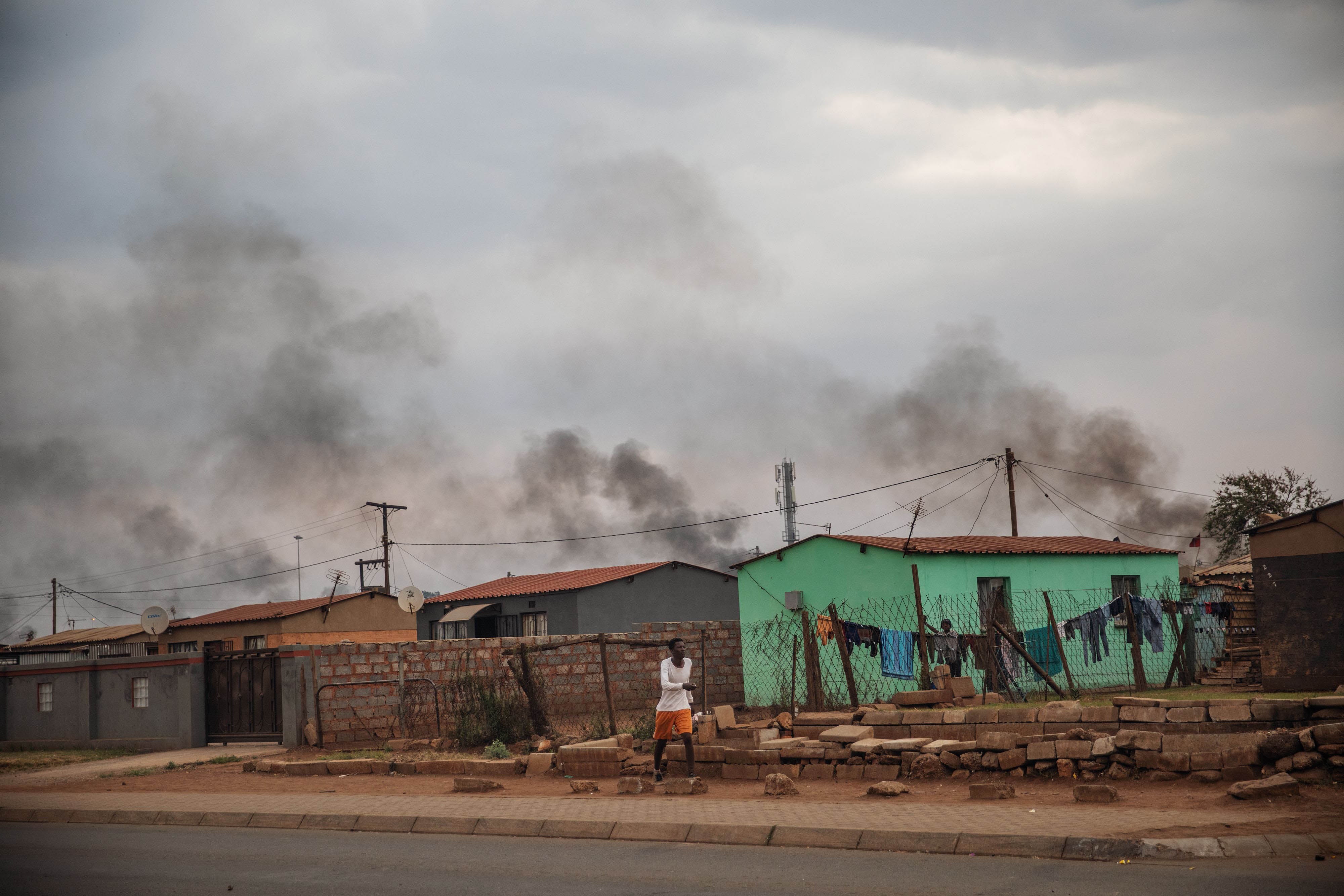 A man walks in front of a buildings, with smoke rising in the background