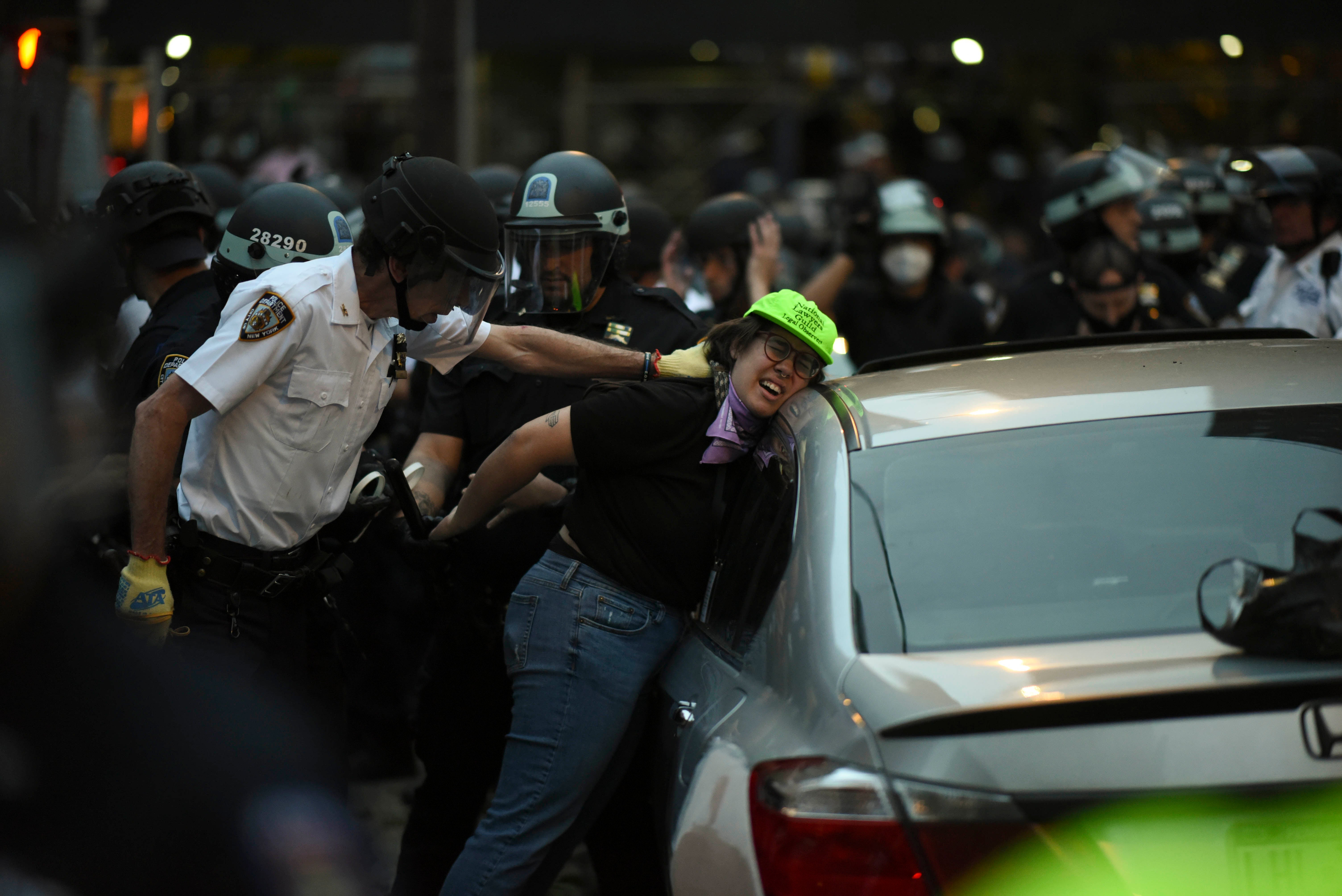 Police push a woman in a green hat against a car and handcuff her