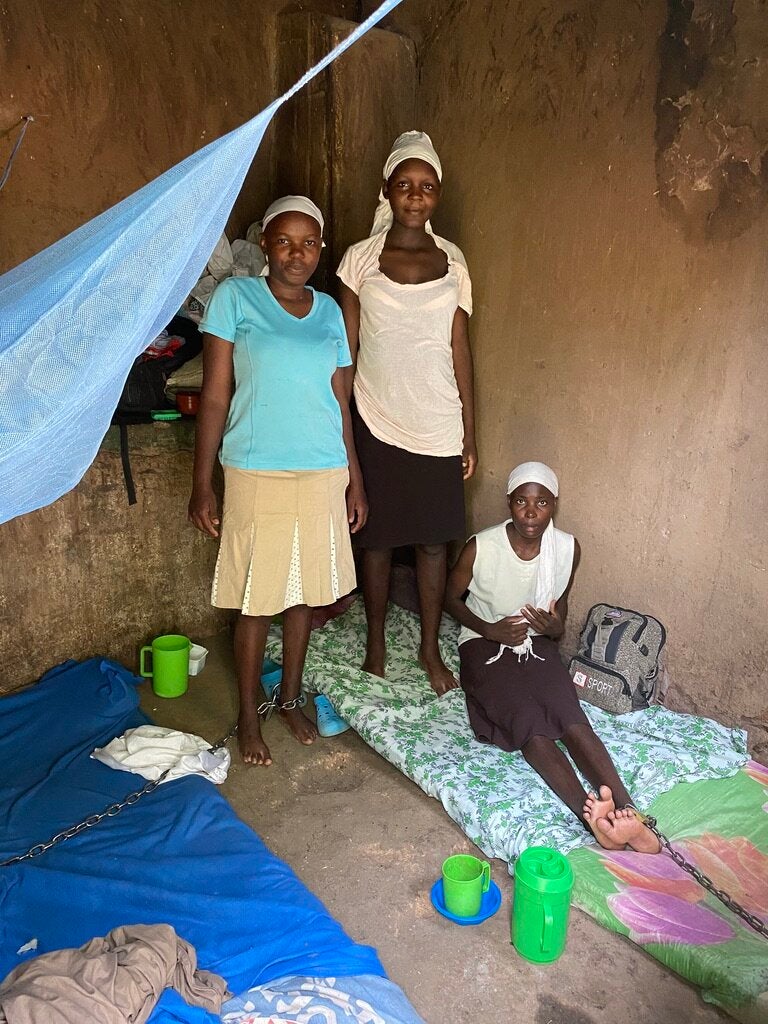 Two women standing and one sitting on the floor in a small room. Two of the women have chains around their ankles.
