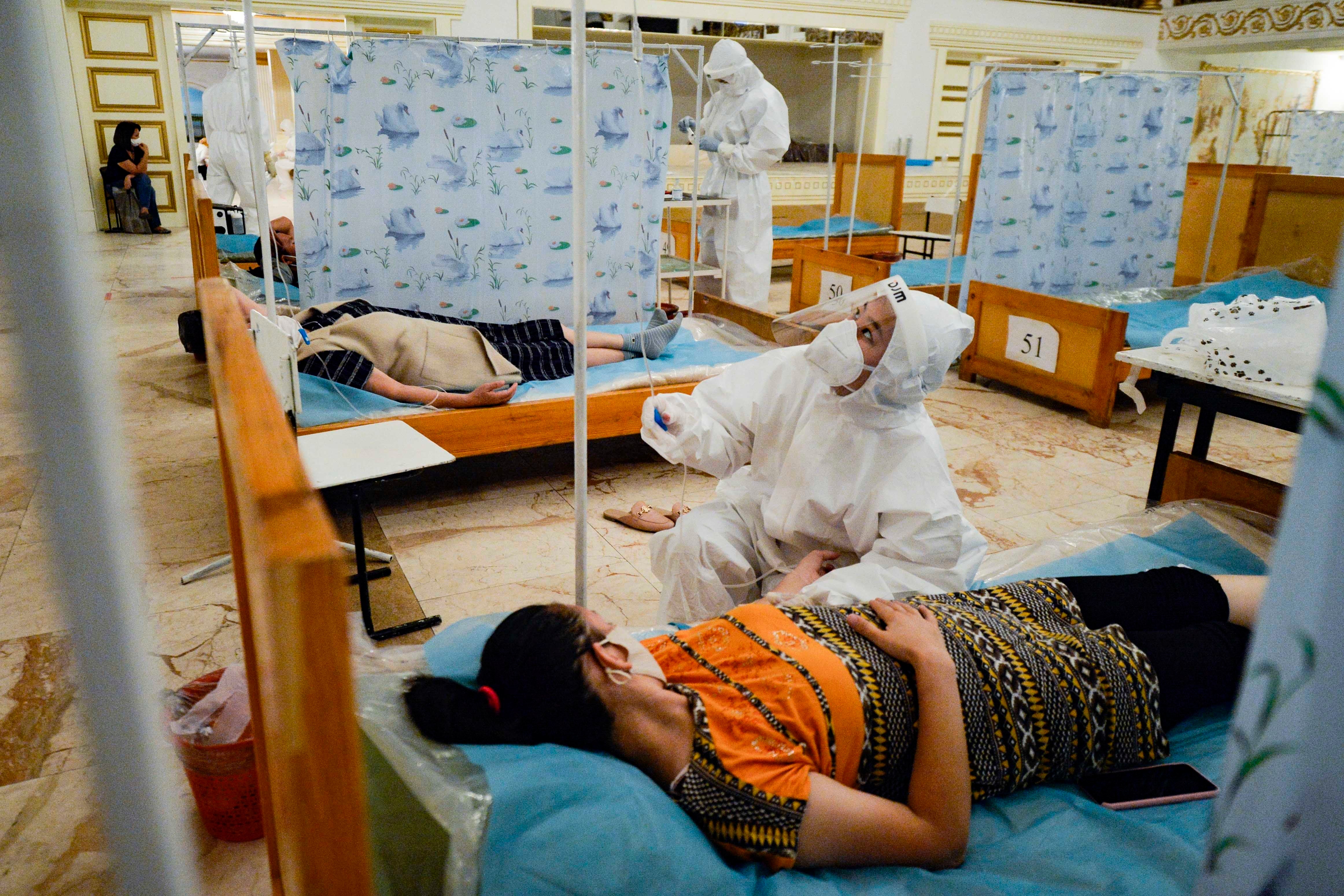 A healthcare worker in a suit and mask checks a patient's IV in a hospital room