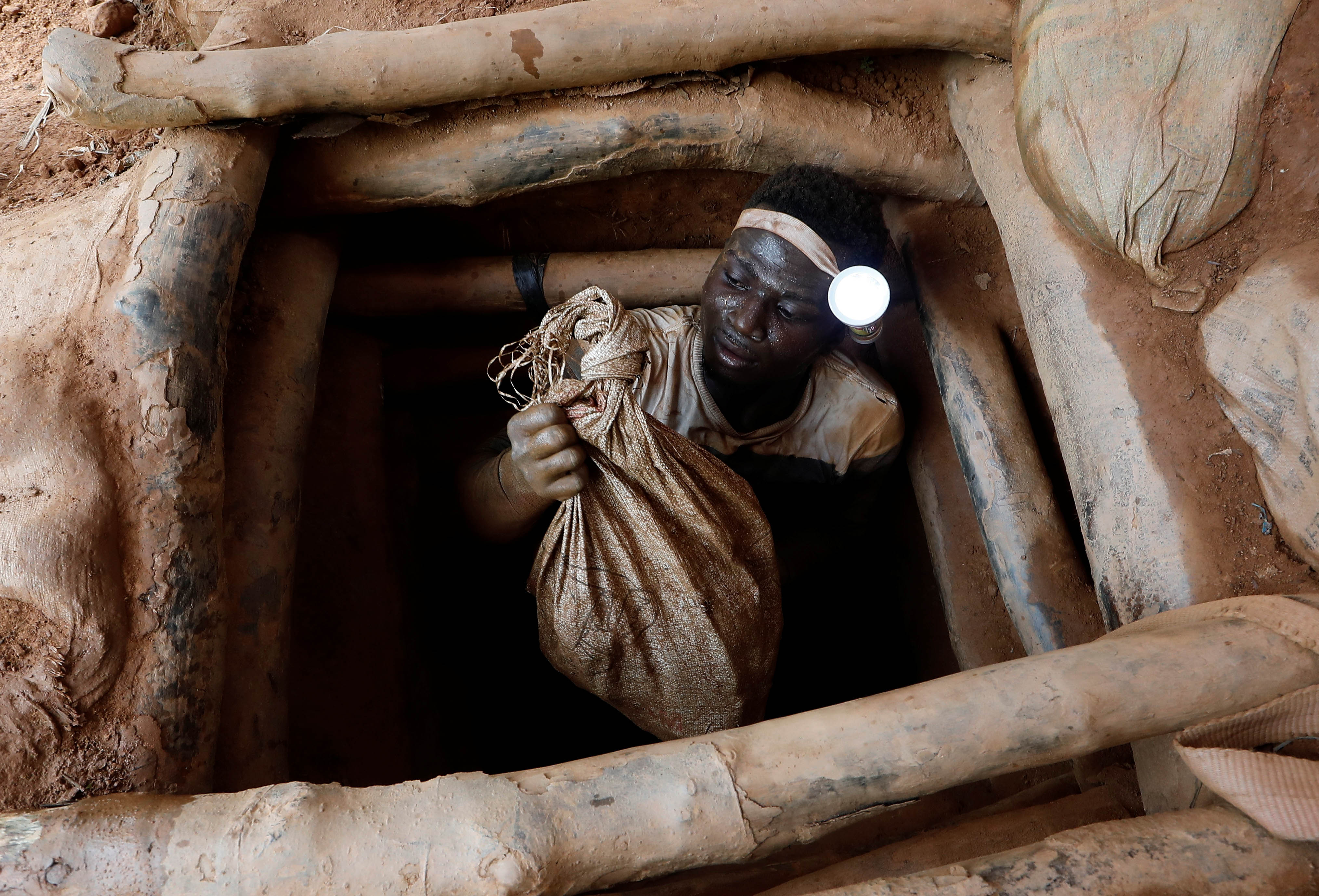 A man wearing a flashlight and holding a bag climbs out of a pit