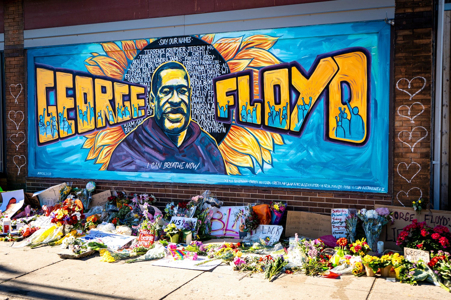 A mural dedicated to George Floyd and honoring the black lives matter movement, showing a depiction of George Floyd's face with his name and supporters of the movement against a blue painted background. Flowers and handwritten signs adorn the sidewalk beneath the mural.