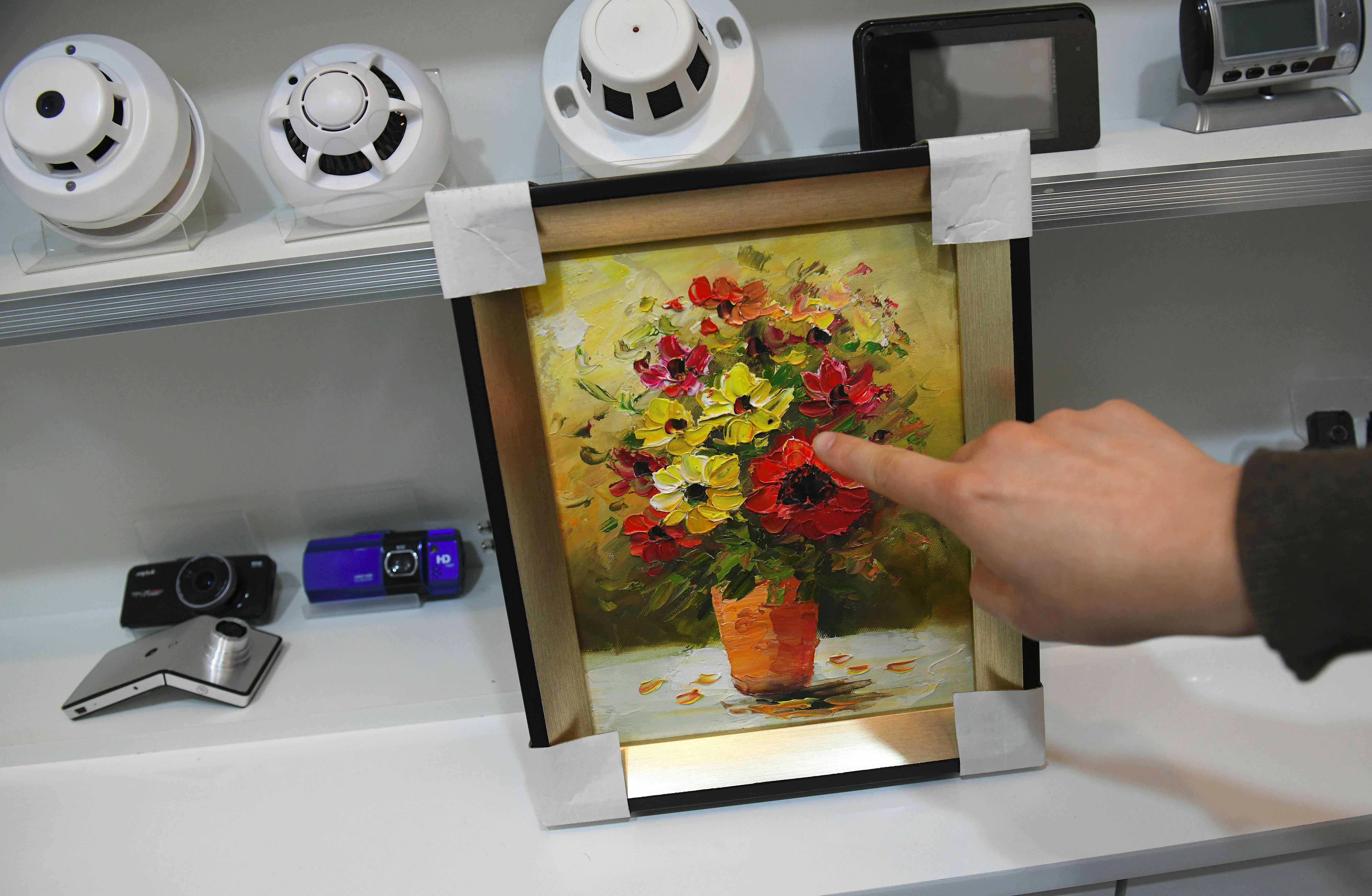 A hand points towards a small painting with a hidden camera inside it 