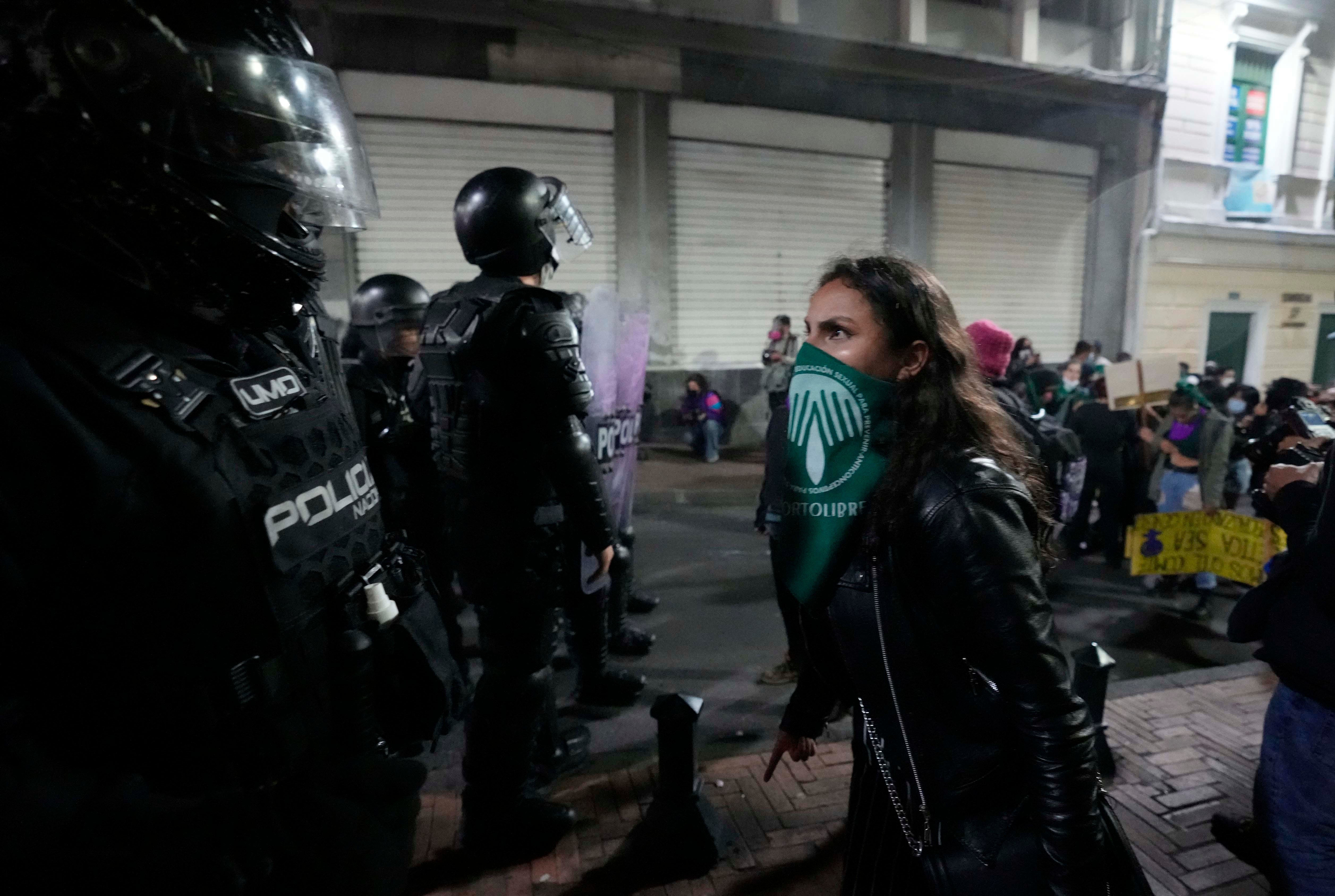 A woman wearing a green bandana over her face stands in front of a police officer with riot gear