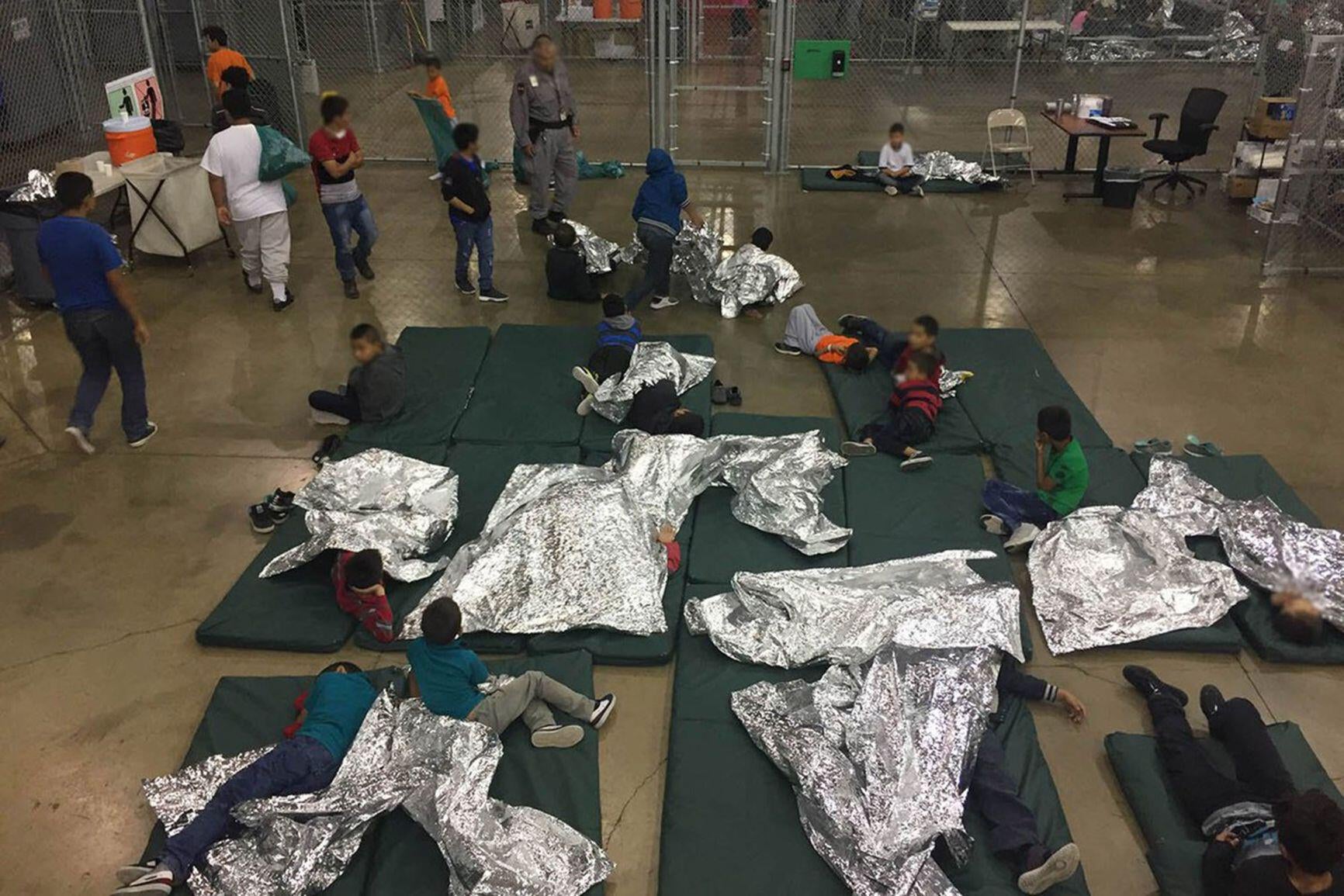 Children in a large indoor fenced-in holding area, wrapped in metallic blankets