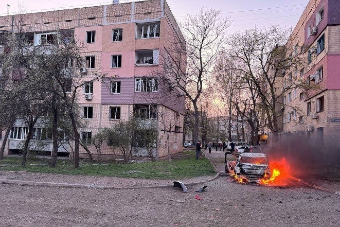 A white car on fire between two apartment buildings damaged by a Russian air-burst munition