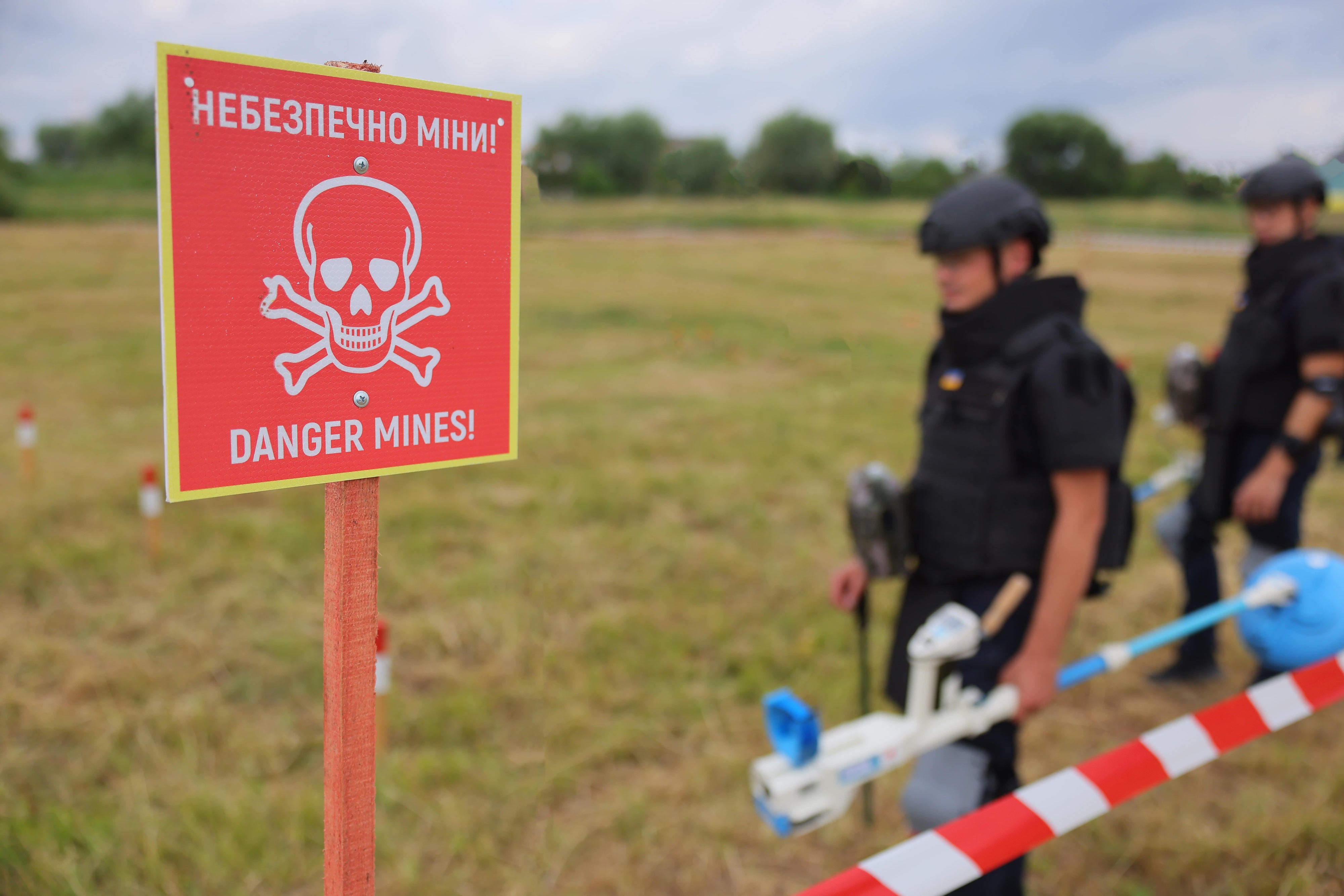 Sign saying "danger mine" in Ukrainian and English with deminers in the background.