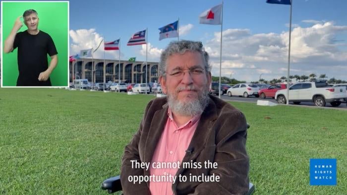 A man looks at the camera while speaking in front of a government building in Brazil. A sign language interpreter is in the top left corner and the HRW logo is in the bottom right.