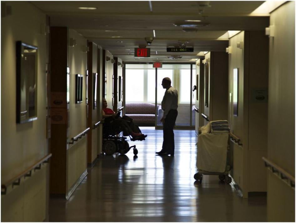 A woman in a wheelchair talks to a man who is standing in a nursing home.