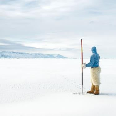 Photograph showing an indigenous man holding a spear and standing on a sea of ice looking out at snow capped mountains.